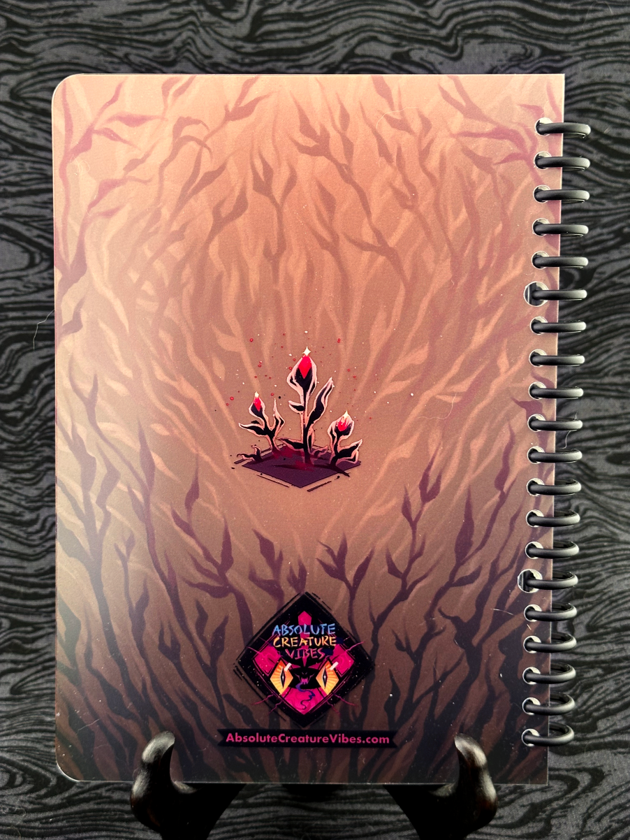 Spiral bound notebook back cover featuring small black plants with pink flower buds on a tan background. Absolute Creature Vibes logo and website url at the bottom.