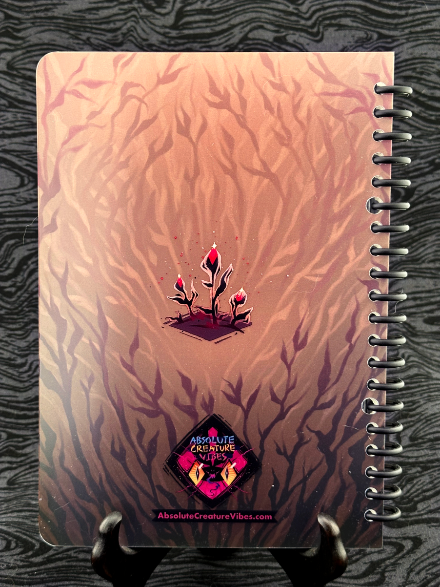 Spiral bound notebook back cover featuring small black plants with pink flower buds on a tan background. Absolute Creature Vibes logo and website url at the bottom. 